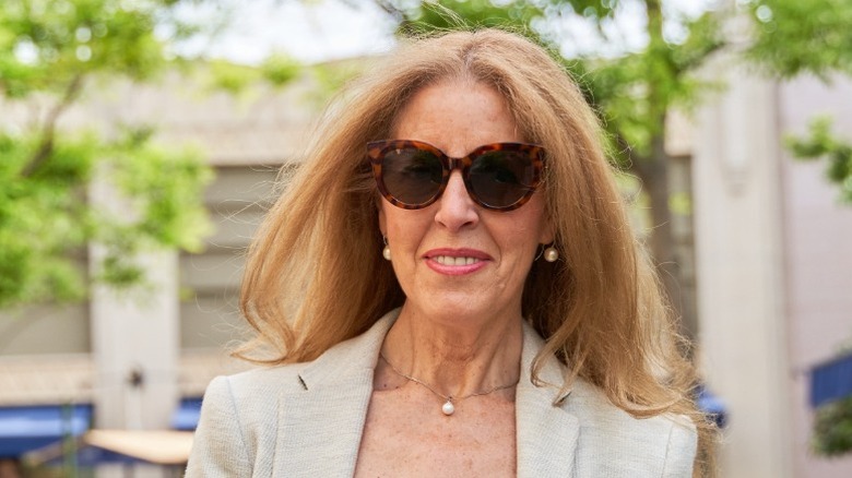 Une femme aux cheveux longs portant un blazer beige, des bijoux en perles et des lunettes de soleil.