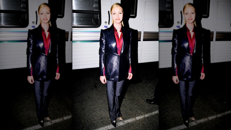 Lisa Kudrow porte une veste en cuir noire avec une chemise rouge et un pantalon gris avec les cheveux lissés en arrière.