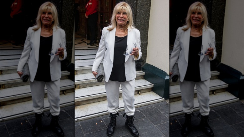 Nancy Sinatra photographiée à l'extérieur tenant ses lunettes et portant un pantalon blanc, un blazer avec une chemise noire et des bottes de combat.