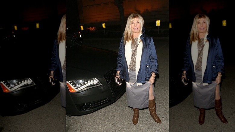 Nancy Sinatra posant lors d'un événement portant une jupe argentée et un haut doré scintillant avec un manteau bleu foncé et des bottes marron.