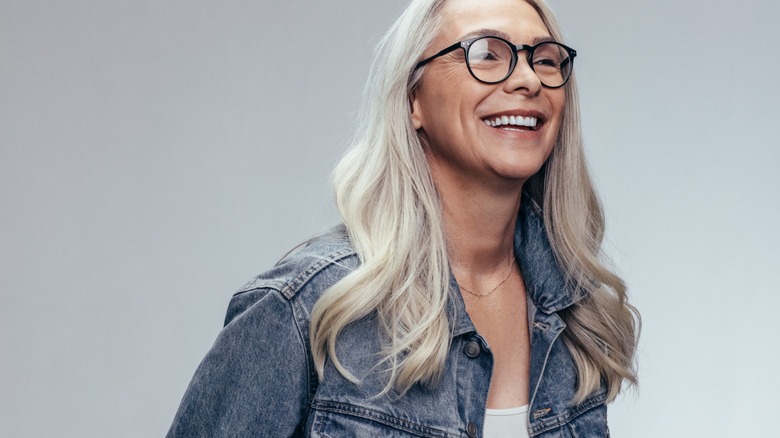 Une femme souriante aux longs cheveux gris portant des lunettes et une veste en jean.