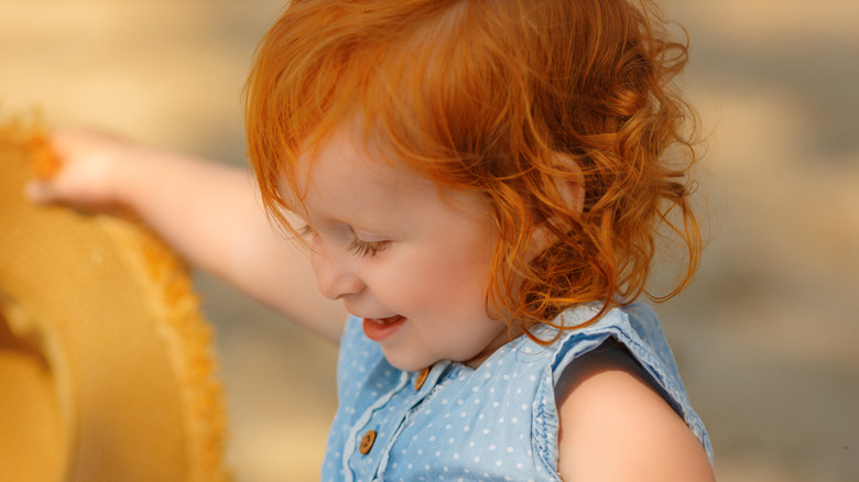 Un tout-petit souriant aux cheveux roux, vêtu d'un haut à pois bleu et blanc, assis dehors.