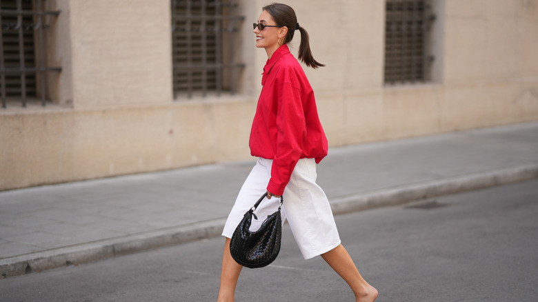 Une femme traverse la rue vêtue d’une veste rouge, d’un short blanc et d’un sac à main noir.