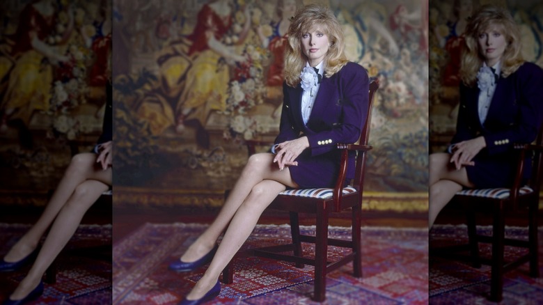 Morgan Fairchild assis sur une chaise, les jambes croisées, vêtu d'un tailleur jupe violet foncé.