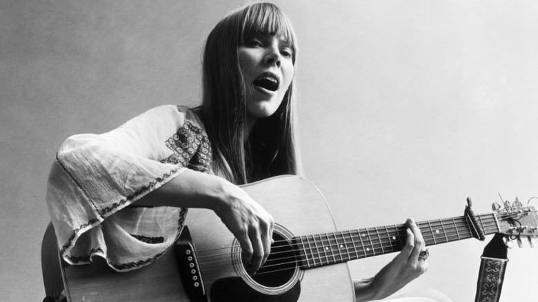 Un portrait de Joni Mitchell assise par terre en train de jouer de la guitare