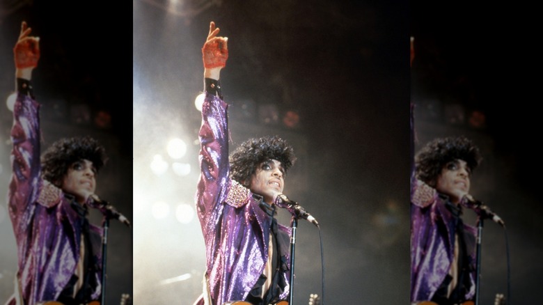 Prince se produisant lors de la tournée Purple Rain portant une veste violette scintillante, la main en l'air.