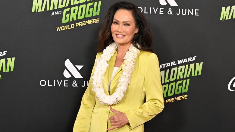 Tia Carrere lance une pancarte shaka lors d'un événement sur le tapis rouge, vêtue d'un costume jaune clair et d'un collier.