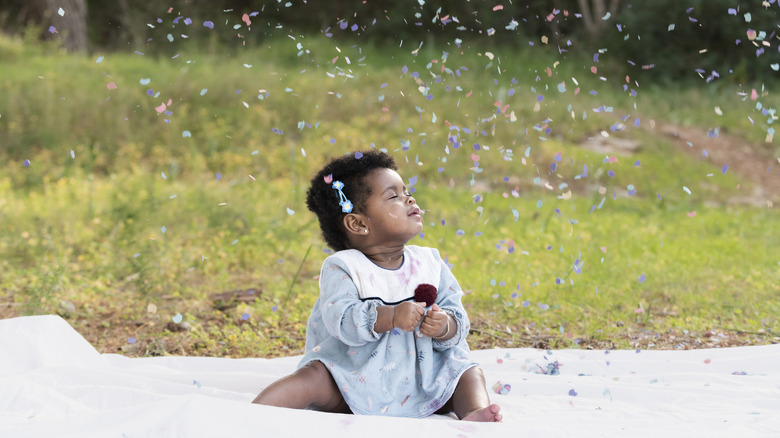 Petite fille assise sur une couverture, souriant les yeux fermés alors que des confettis colorés tombent autour d'elle
