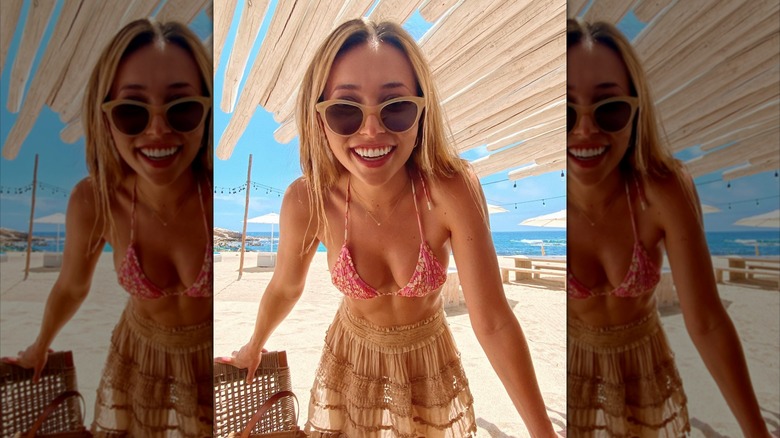 Camelia Somers porte des lunettes de soleil, un haut de bikini à fleurs rose et une jupe transparente marron clair sur la plage.