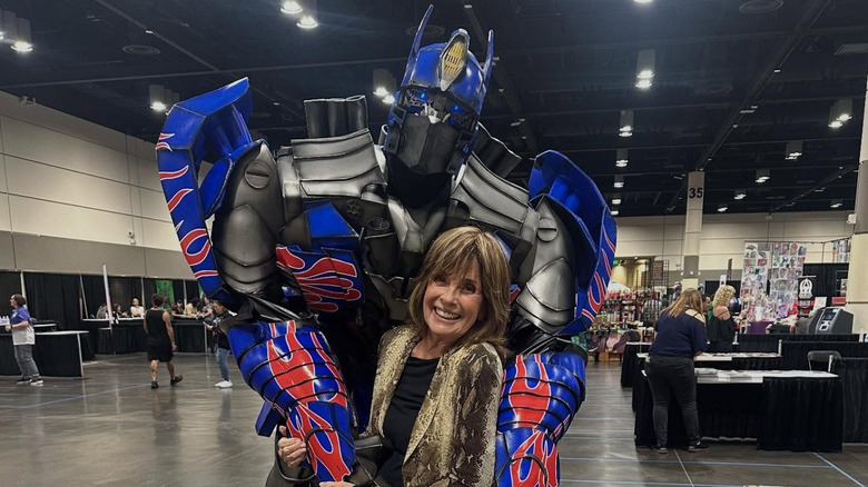 Linda Gray pose avec quelqu'un vêtu d'un costume de Transformers dans une salle événementielle